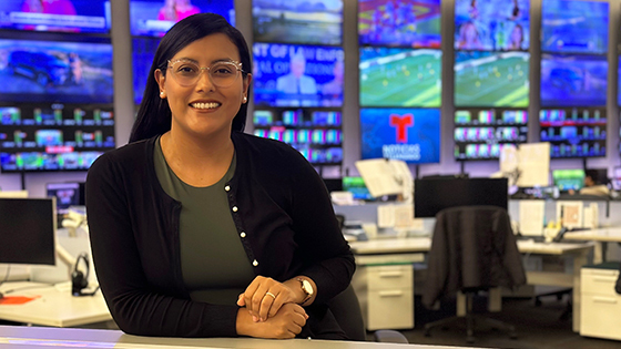 Priscilla sits at a desk in a live news television studio. She is wearing glasses and is smiling.