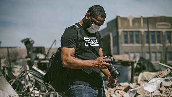 Grad E Mackey looking down at a DSLR camera while wearing a grey face mask, backpack, and black shirt with the word "press" in white, standing amongst a large amount of debris.