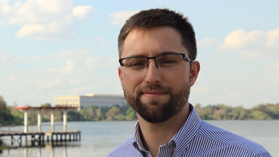 A man wearing glasses and a button-up shirt stands in front of a pier.