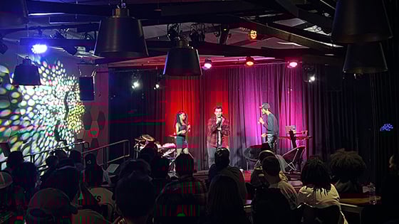 Three performers sing on a stage in front of an audience. Red and pink lights shine behind them.