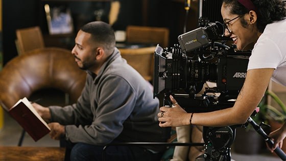 Behind the scenes on a student film set, one student sits in a chair with a notebook while another student works behind a film camera