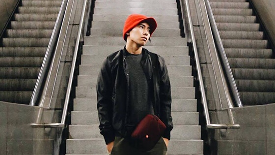 A young Asian man wearing a red knit cap, black jacket and shirt, green pants, and a burgundy bag stands between two escalators. His hands are in his pockets and he is looking off to the left.