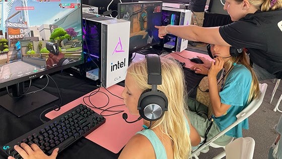 Two young girls are seated at desktop computers playing 'Minecraft' while a woman wearing a Super Girl Gamer Pro shirt assists them.