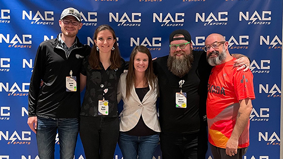 Armada graduates Nathan Duke and Megan Danaher and Full Sail esports staffers Sari Kitelyn, Bennett Newsome, and Jacob Kaplan stand in front of a NACE backdrop.