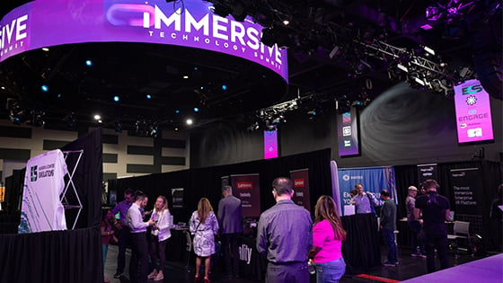 A large room with lots of booths set up, each from a different technology company. People in business-casual clothing are walking around and socializing.