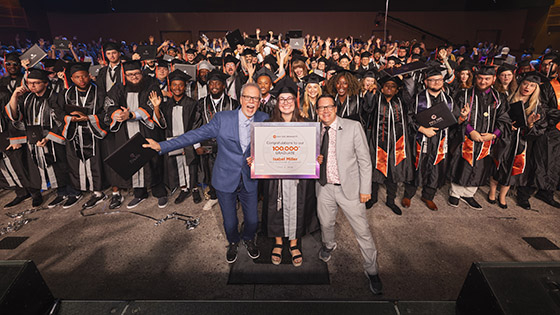 In a large venue full of college graduates, two men in suits stand on opposite sides of a woman in a cap and gown holding a framed photo that reads "Full Sail University - Congratulations to our 100,000th graduate Isabel Miller."