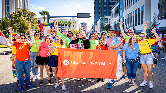 A group of Full Sail faculty, students, and staff posing with a large orange banner that reads “Full Sail University”