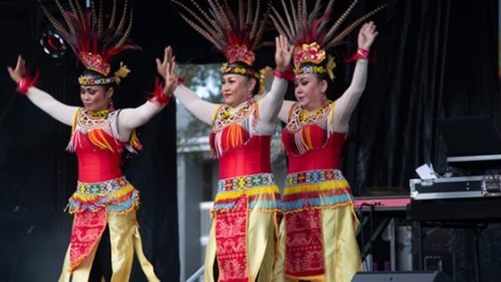 Three women dancing in colorful Indonesian feathered headdresses, yellow skirts, and beaded belts and collars.