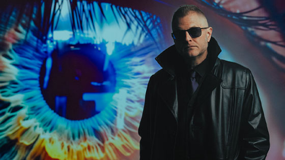 A man in a black leather coat and sunglasses stands before a giant, colorful close-up of an eye glowing with blue and orange light.