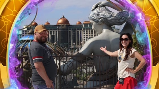 A man and woman stand together in front of an attraction featuring a large statue of a woman, a building with a glass dome, and a building with bronze domes.