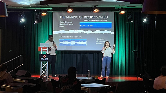 Tyonne Heyward and Ivana Loyola stand onstage in front of an audience. They share a slideshow presentation on the screen behind them.
