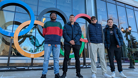 Aca Anderson, Johnathan Richardson, Jackson Faulknor, and Josh Minker stand in a row in front of the NBC Sports building with the colorful Olympic rings and a large sign displaying “NBC Sports.”
