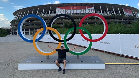 Grad Andrew Molina sits outdoors with his arms spread in front of a set of Olympic rings. There is a stadium with a banner for the 2020 Tokyo Olympics behind him.