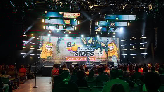 A large audience sits in chairs in front of a stage, which has a digital backdrop that says 2024 B-Side Orlando Chomp Your Own Adventure with two cartoon alligators.