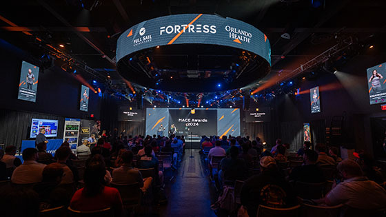A large audience sits in Full Sail’s esports venue. A stage with a few people standing on it is at the front of the room. The words NACE Awards 2024 are on the wall behind them.