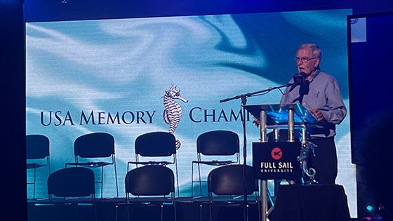 A man wearing glasses and a collared shirt speaks at a podium on a stage. A screen that says USA Memory Championship is behind him.