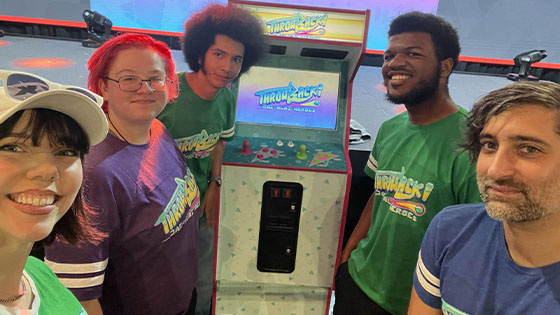 Caris Baker stands with four students in front of an arcade game machine that says Throwback! Jai-Alai Heroes on the screen.