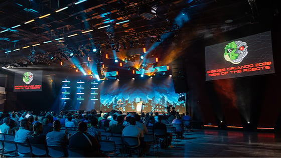 Attendees sit in the Full Sail Live venue. Screens with a lizard robot logo and the words BSides Orlando 2023: Rise of the Robots are on either side of the stage.