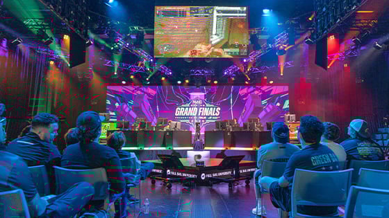 An audience faces a stage where eight competitors sit at consoles. A screen above them shows the game they're playing and another screen displays the NACE Grand Finals 2025 graphic.