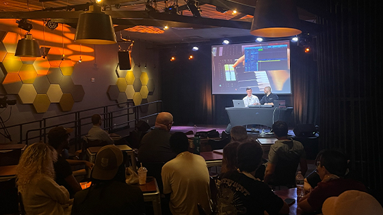 Scott Dickinson and Evan Weiss sit onstage with a keyboard and two laptops. Students sit in an audience facing the stage.