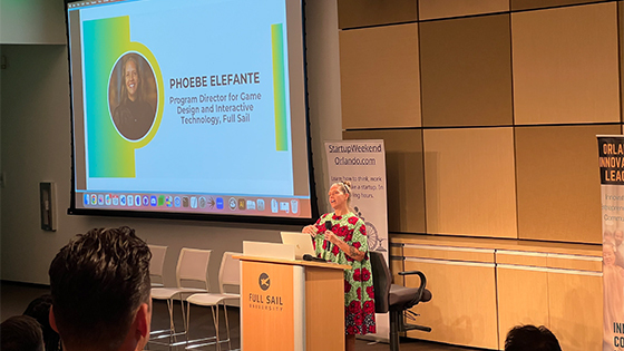 Phoebe Elefante stands at the podium speaking to the audience in the Full Sail venue. She is wearing a brightly colored print dress.