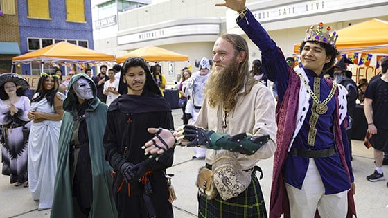 Several students dressed in costumes including a mushroom, a goddess, a viking, and a king stand in a line. Orange tents surround the crowd of students.