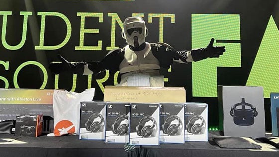 A masked figure stands behind a table piled with boxes of headphones and various technology on stage at the Student Resource Fair.