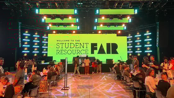 Students and staff sit at tables and interact. A large screen behind them displays “Welcome to the Student Resource Fair” in black text with a green background.