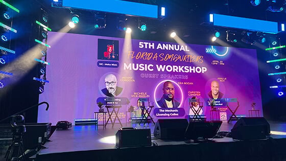A large screen reads "5th Annual Songwriters Music Workshop" against a purple backdrop. The screen sits atop a brightly lit, empty stage.