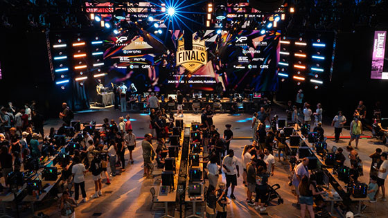 Four rows of 12 gaming consoles each fill a large room. Bystanders watch as competitors play various games at their stations. A large screen displays an XP League North American Finals graphic.