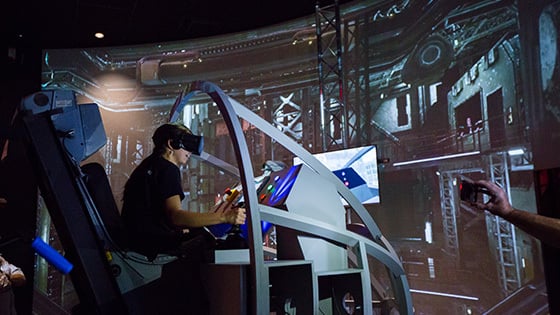 A student wearing a VR headset sits in a space shuttle landing simulator. A large screen displaying a warehouse is in front of them.