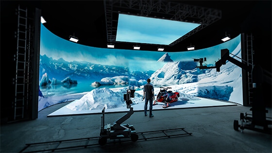 A man wearing jeans, sneakers, and a plaid shirt stands in a production studio. He is facing a large LED screen showing an icy mountain range and a snowmobile.