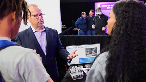 Man wearing glasses and a blue suit speaks to guests at Career Fair while man in back tries on Virtual Reality goggles.