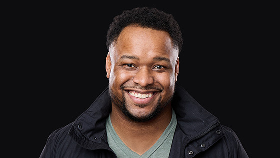 James Lewis smiles at the camera. He is wearing a black jacket and green v-neck t-shirt.