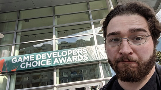 Ryan Miller pictured from above the shoulders in a black leather jacket with silver wireframe glasses in front of a sign reading “Game Developers Choice Awards.”