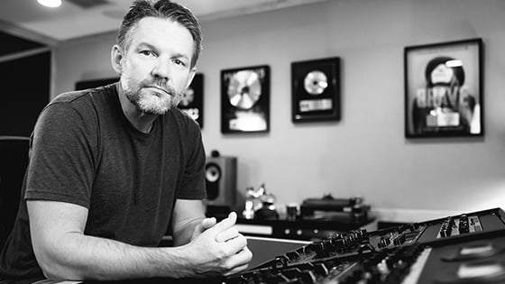 Brad Blackwood sits at an audio console in a studio with framed gold records on the wall behind him.