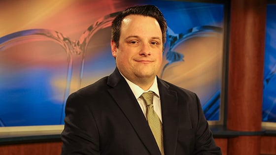 Dan Hanson sits at a news desk in a television studio. He is smiling and wearing a black suit with a yellow tie.