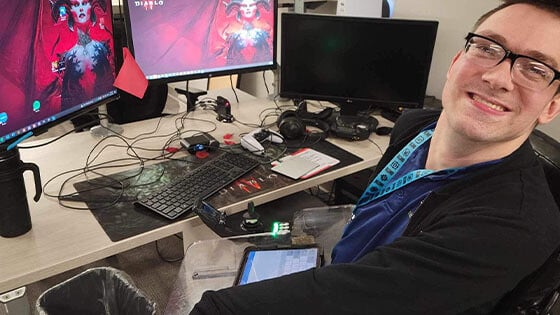 Brock Davis sits at his desk at Blizzard Entertainment. Two computer screens displaying the ‘Diablo IV’ logo are on the desk in front of him.
