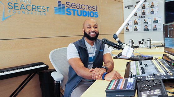 Full Sail grad Anthony Grier sits in front of an audio board at Orlando Health Arnold Palmer Hospital for Children’s Seacrest Studios, smiling.