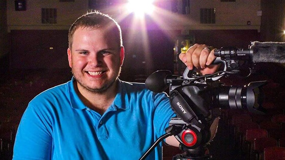 Grad Cody Grammer in a blue collared shirt, holding a video camera in a movie theater.