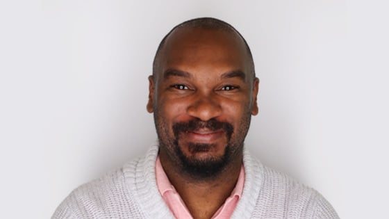 Quentin stands in front of a white wall wearing a white cardigan over a pink polo. His arms are crossed and he is smiling.