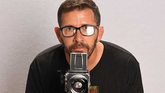 Sebastian Krys crouches behind a video camera. He is wearing black-rimmed eyeglasses and a black t-shirt with a logo on it.