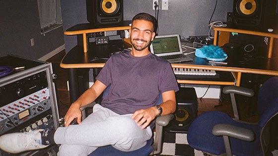 A smiling man is seated casually in a music studio, surrounded by audio equipment, a keyboard, speakers, and a laptop on a desk.