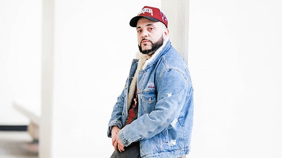 A man wearing a red baseball cap and a denim jacket is pictured leaning against a concrete pillar in a brightly lit room.