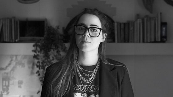 Veronica Concha stands in front of a bookshelf in her home. She is wearing a dark blazer, a band t-shirt, silver necklaces, and glasses.