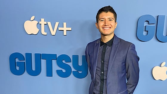 A person with short, dark hair is smiling while wearing a blue suit against a light blue step and repeat that shows the logos for ‘Gutsy’ and Apple TV+.