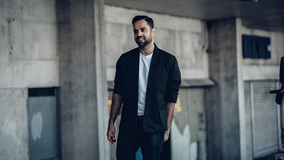 A well-dressed man in a white t-shirt and black blazer posing in front of a worn-down building.