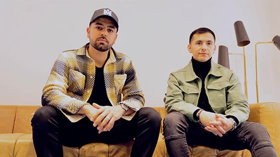 Two men seated side by side on a light brown leather couch against a white backdrop.