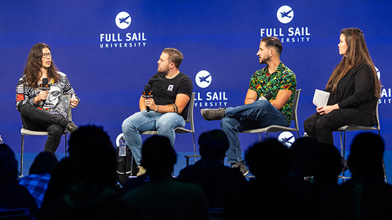 Four people seated on a stage, talking and holding microphones in front of a large crowd.