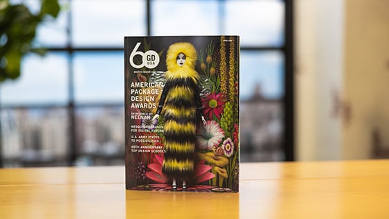 Graphic Design USA’s April 2023 issue sits upright on a wooden table. The cover features a female doll wearing a long furry black-and-yellow dress with a hood. She is standing in front of a psychedelic floral background.